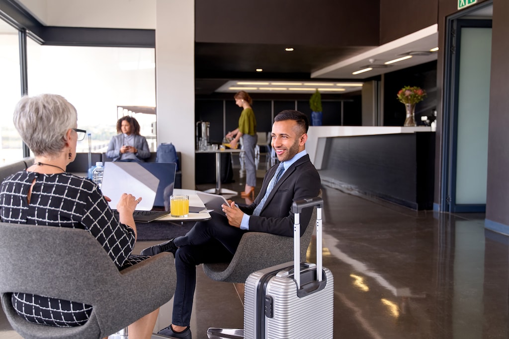 Two mixed race positive coworkers communicating brainstorming ideas while working on a laptop and tablet in modern airport, waiting room during business trip with carry-on luggage