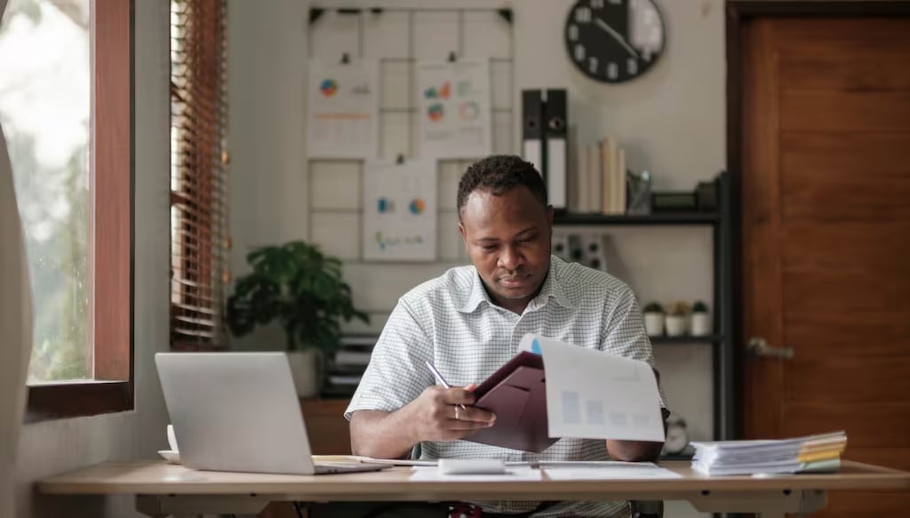 A photo of a man calculating net income while reviewing financial documents at a desk.