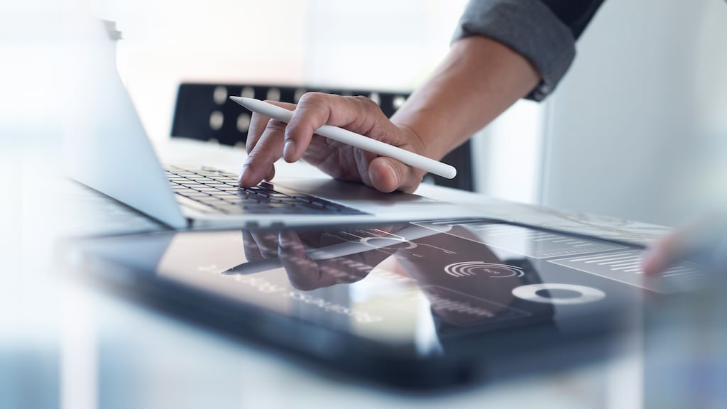 Eine Person arbeitet an einem Laptop und analysiert gleichzeitig einen Marktbericht auf einem Tablet, das auf dem Tisch liegt. 