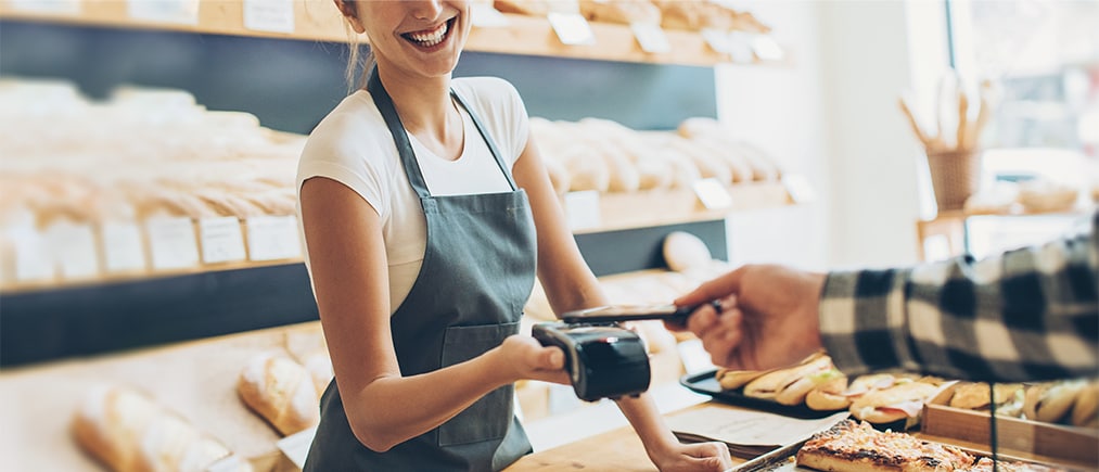 Mitarbeiterin einer Bäckerei steht lächelnd an der Kasse und reicht einem Kunden ein EC-Kartenlesegerät. 
