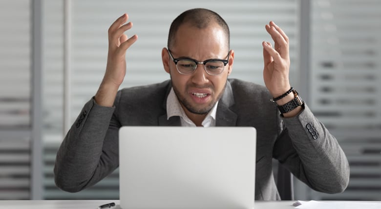 Ein Mann sitzt mit erhobenen Händen vor seinem Laptop und ärgert sich.