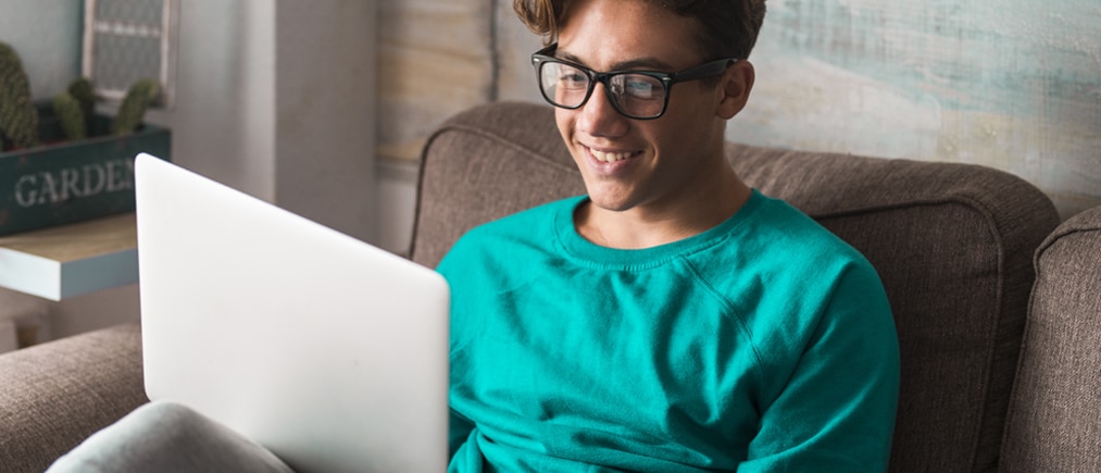 Junge Person sitzt lächelnd vor Laptop auf einer Wohnzimmercouch