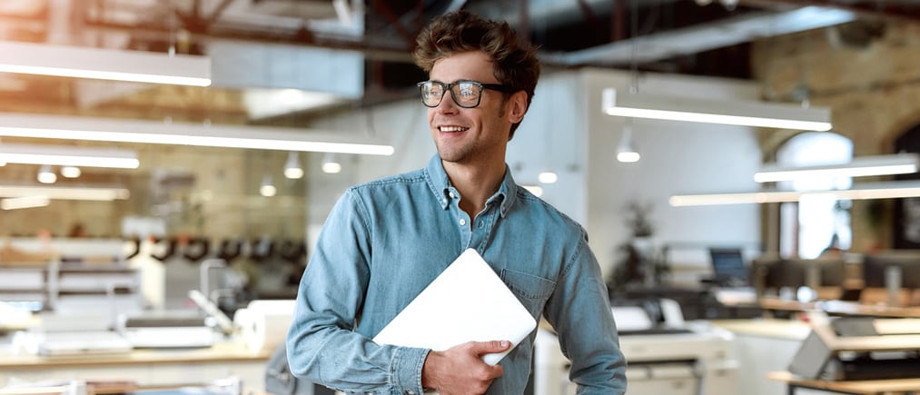 Junger Geschäftsmann mit Brille und Laptop in der Hand posiert inmitten eines Produktionsbetriebes.