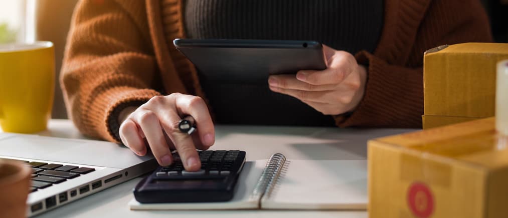 Eine Frau sitzt an ihrem Schreibtisch und hat dabei einen Taschenrechner und ein Tablet in der Hand. 
