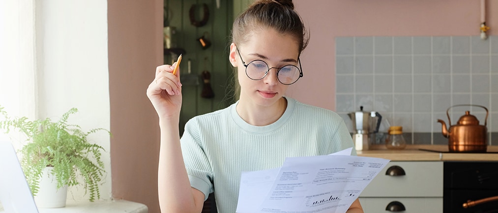 Indoor picture of young European female sitting at home at table reading data in sheets of paper with pencil in order to figure out something important and check it, feeling relaxed and confident