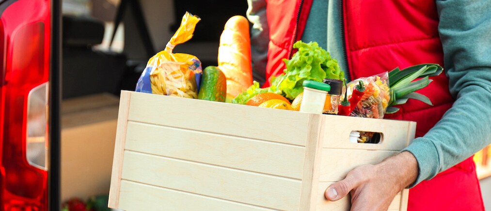 Courier holding crate with products near car outdoors, closeup. Food delivery service