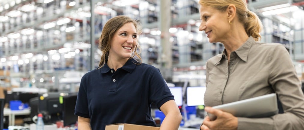 Female worker holding packages talking with warehouse manager and smiling. Large distribution company employees walking together and talking.
