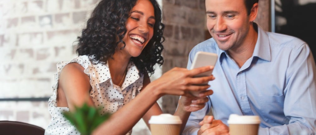 Businessman and businesswoman looking at a mobile phone. Multi ethnic group with African American and Caucasian. They are casually dressed and having a discussion. They are drinking coffee, laughing, smiling and having fun. Could be reading or sending a text message.