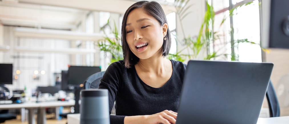 Asian businesswoman talking to virtual assistant at her desk. Female professional working on laptop and talking into a speaker.