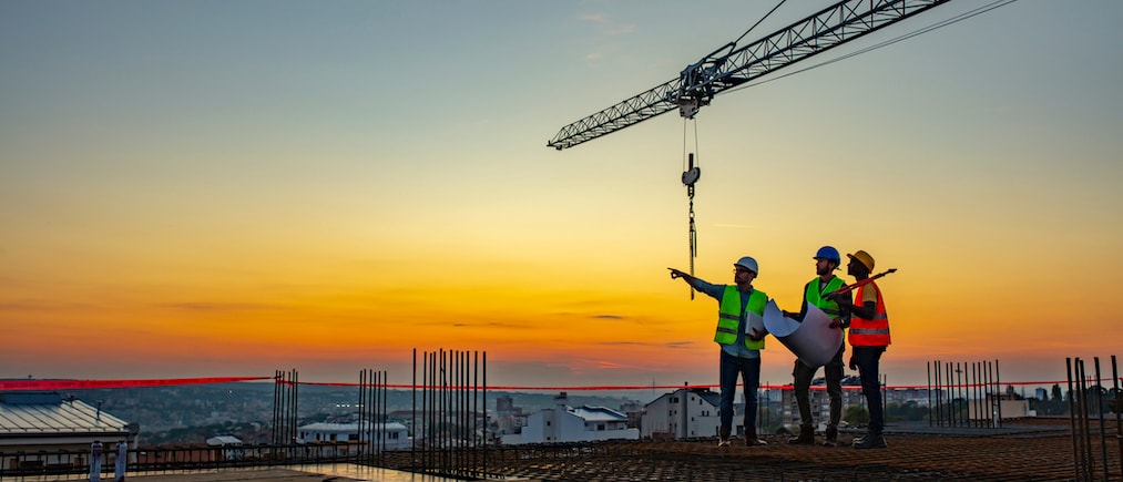 Three Multi-Ethnic construction workers in uniform standing at construction site with crane in background, discussing building plans while holding blueprint at sunset under the tower crane. (Three Multi-Ethnic construction workers in uniform standing 