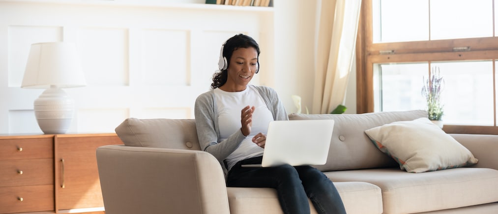 Young African American woman in modern headphones sit on couch in living room talk on video call on laptop, millennial biracial female in earphones relax on sofa at home watch webinar on computer