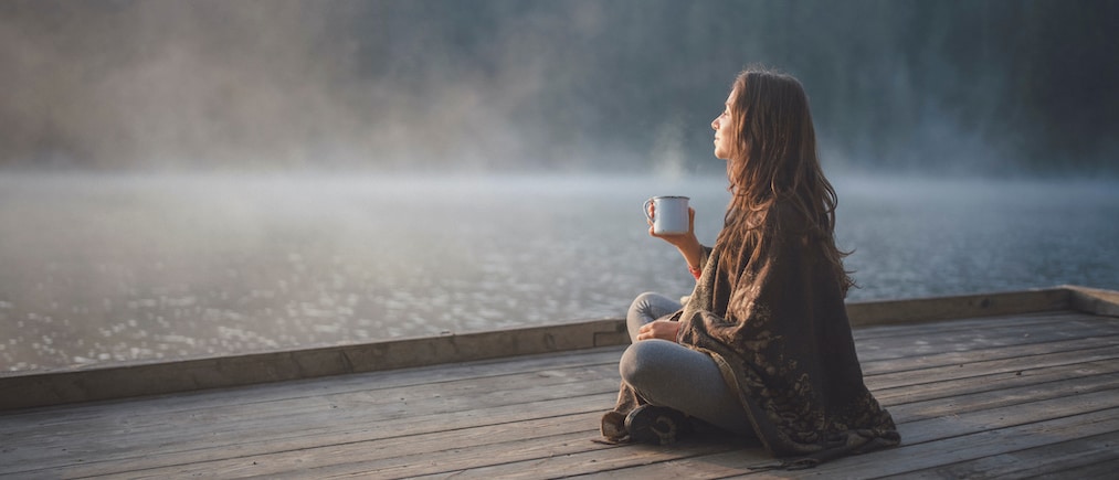 Side view of woman sitting on wooden pier in nature, she looking the sunrise and drinking coffee.