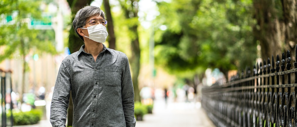 Mature gray-haired Taiwanese man with surgical mask walking on a sidewalk near public park