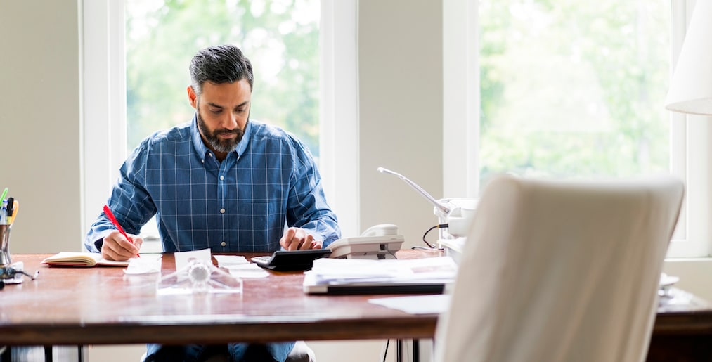 Mid adult businessman using calculator while writing in diary at home office. Serious male is calculating at desk. He is working at home.