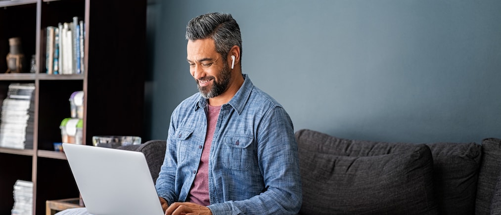 Successful mature indian businessman sitting on couch typing on laptop with wireless earphones. Mixed race businessman sitting on couch while working from home during video call. Happy multiethnic business man in casual clothing working on computer. 
