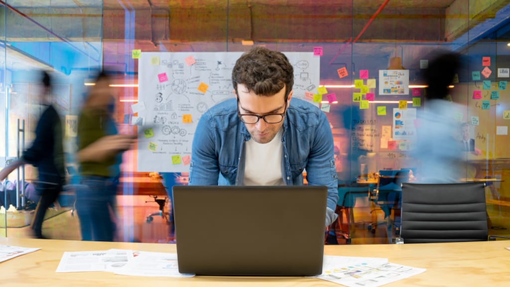 Latin American man working at a creative office using his computer and people moving at the background - place of work concepts 
