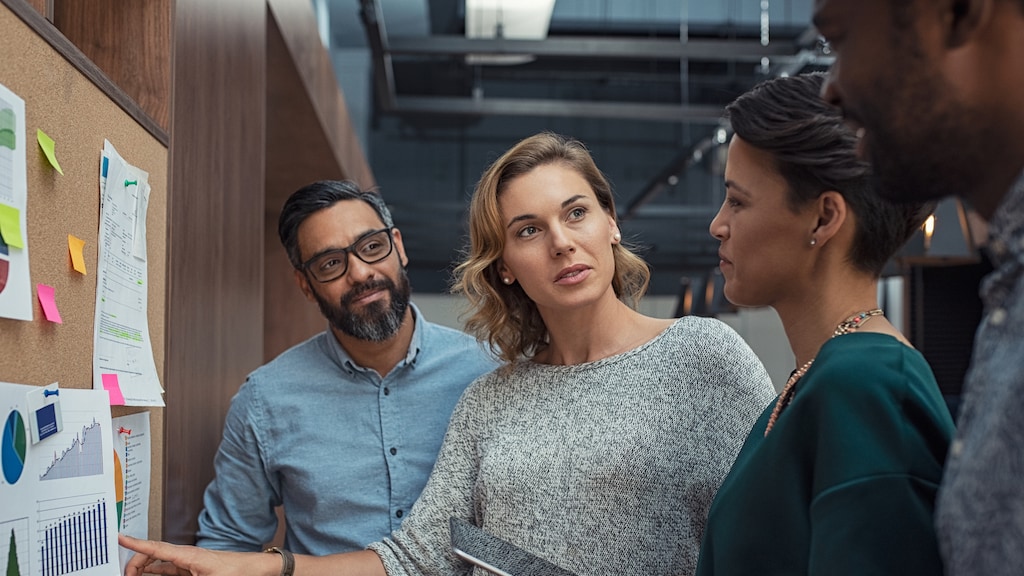 Multiethnic business people discussing charts and graphs showing the results of their successful teamwork. Mature business woman showing presentation to casual colleagues in modern office during night meeting. Group of creatives analyzing market trends.