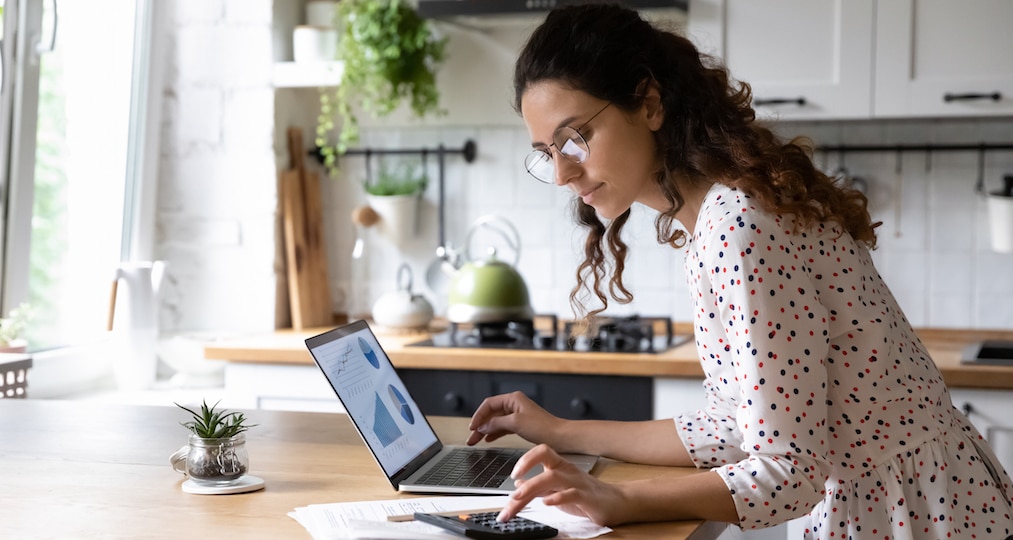 Serious young woman wearing glasses calculating finances, household expenses, confident businesswoman working with project statistics, using laptop and calculator, standing in kitchen at home