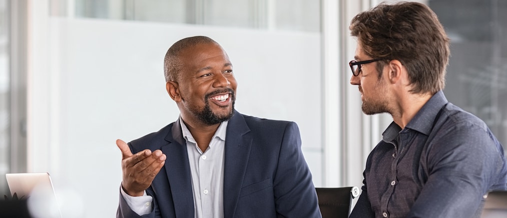Two happy business colleagues at meeting in modern office interior. Successful african boss in a conversation with young employee in boardroom. Marketing team of two businessmen discussing strategy in meeting room.