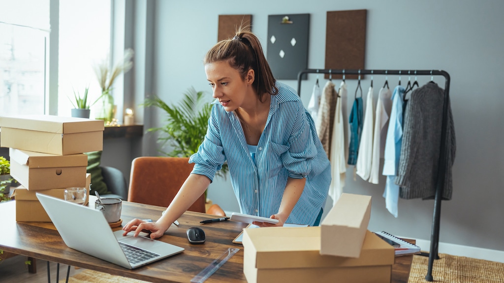 Women, owener of small business packing product in boxes, preparing it for delivery. Working woman at online shop. She wearing casual clothing and checking on laptop address of customer and package information