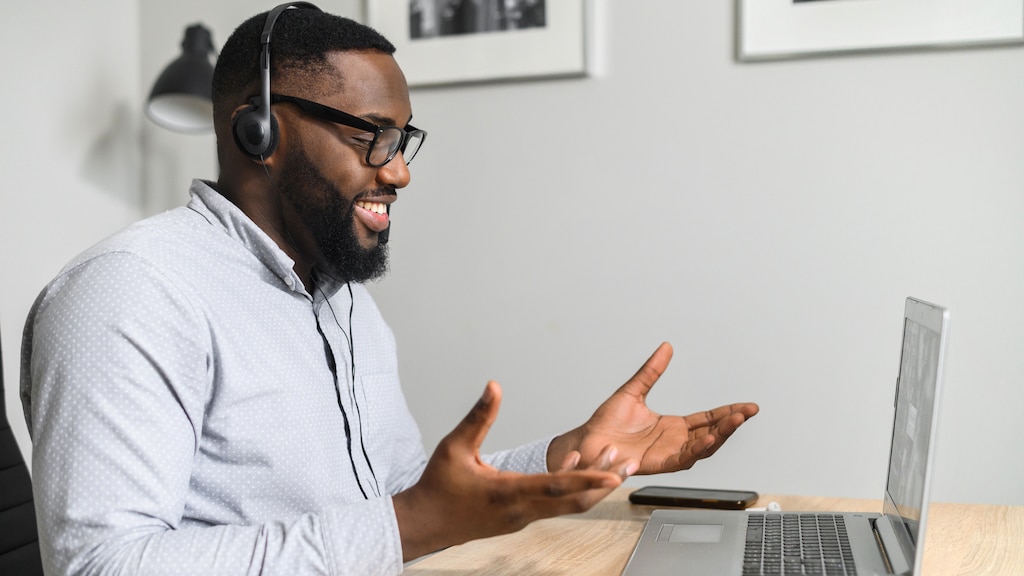 Young intelligent African-American teacher sitting at the desk and having coaching session online with students from business school, working on a laptop, using headphones with a microphone