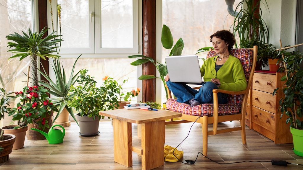 Woman working from home. She is sitting on armchair on her patio surrounded with lot of potted plants and using laptop.