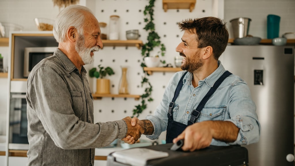 A young handyman shakes hands with an older client, standing in a kitchen
