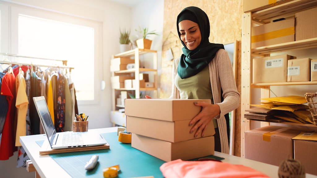 Smiling Muslim woman with hijab packing product into a cardboard box and preparing it for a delivery to a customer. Small business owner.