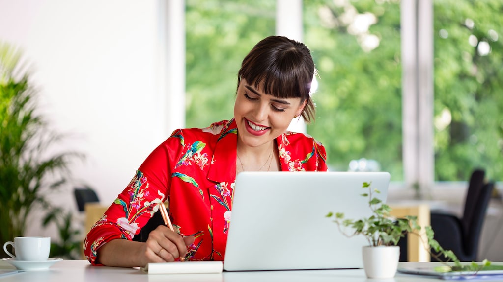 Happy young woman wearing red floral pattern jacket sitting at the desk over laptop in the creative workplace.