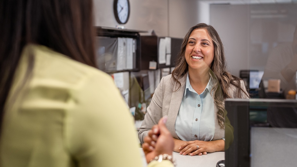 A friendly female customer service representative of Latin American descent greets a customer with a big smile at the counter in a bank.