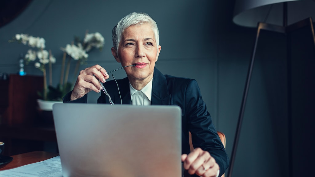 Portrait of mature businesswoman, sitting in her office and thinking. Holding eyeglasses in her mouth. In front of her on the desk is laptop.