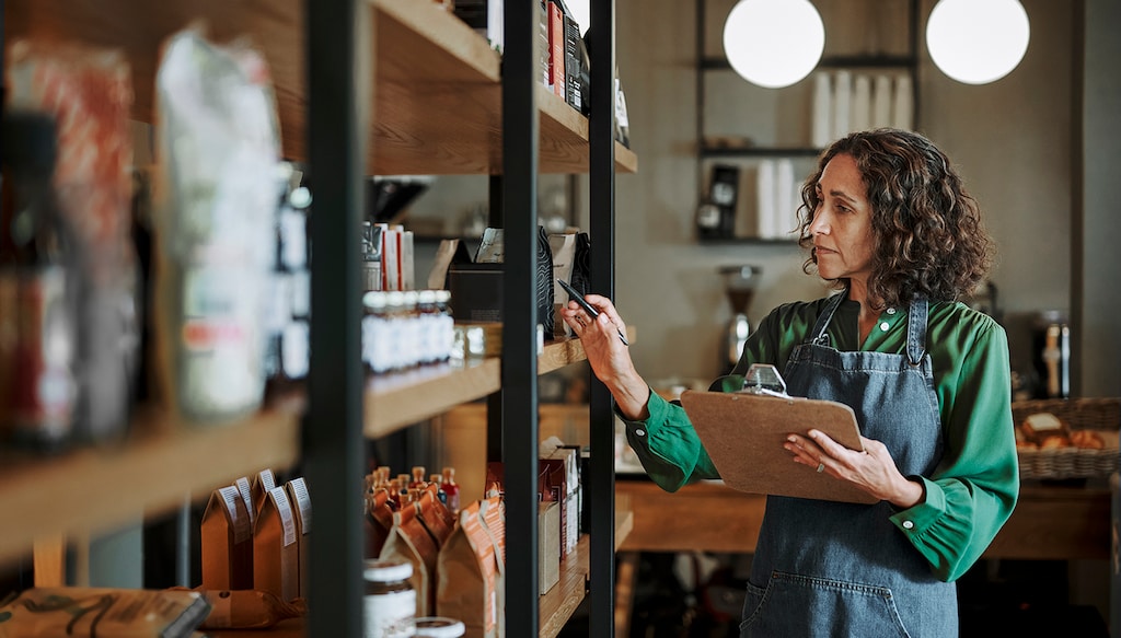 Mature female delicatessen owner holding a clipboard while checking inventory of artisanal merchandise on some shelves in her shop