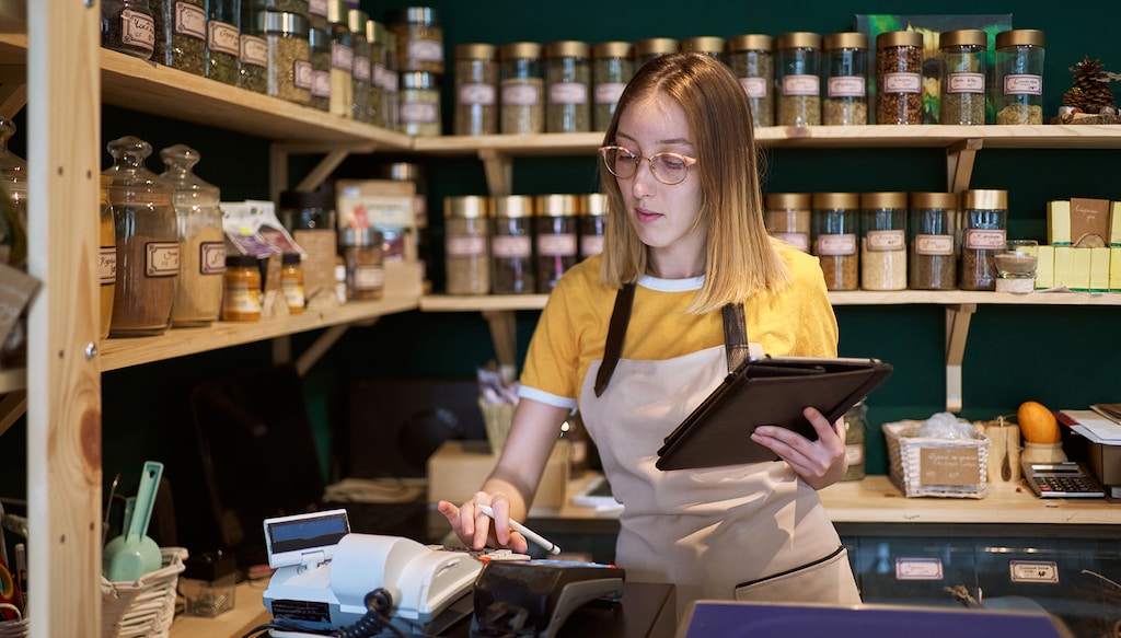 Young female employee at the health food store, organizing products, doing inventory check up and organizing the online orders from the shop website