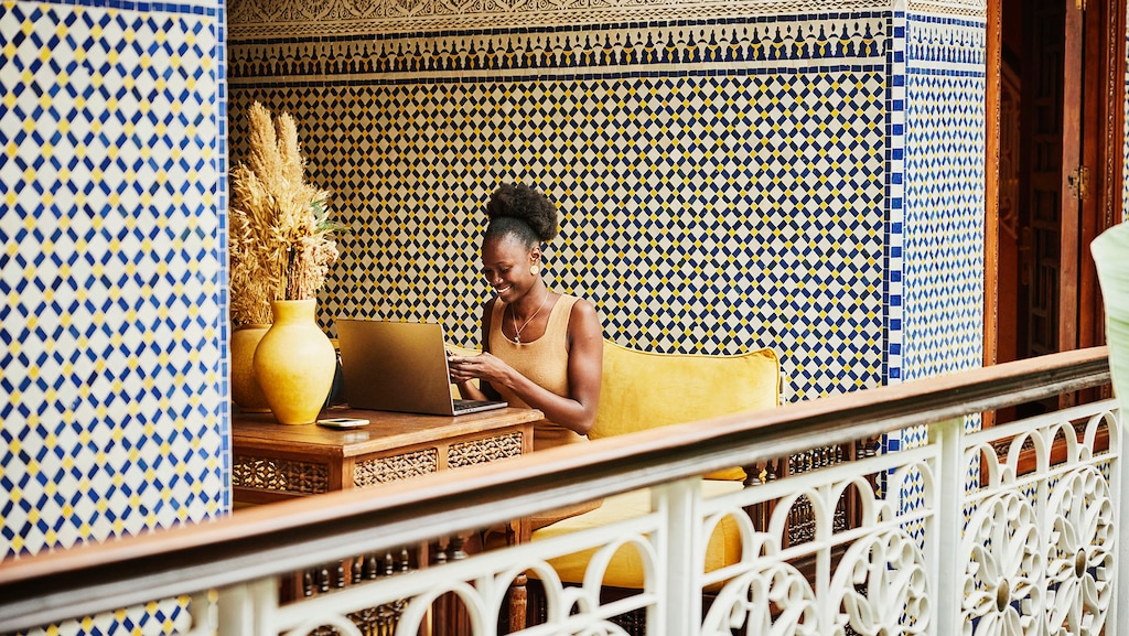 Wide shot of smiling woman checking smart phone working on laptop in alcove of ornately decorated riad while on vacation in Marrakech