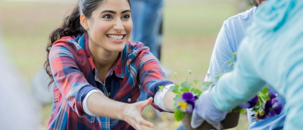A smiling young woman sits on the ground and reaches for some pansies to plant.  She is participating in a community cleanup event.