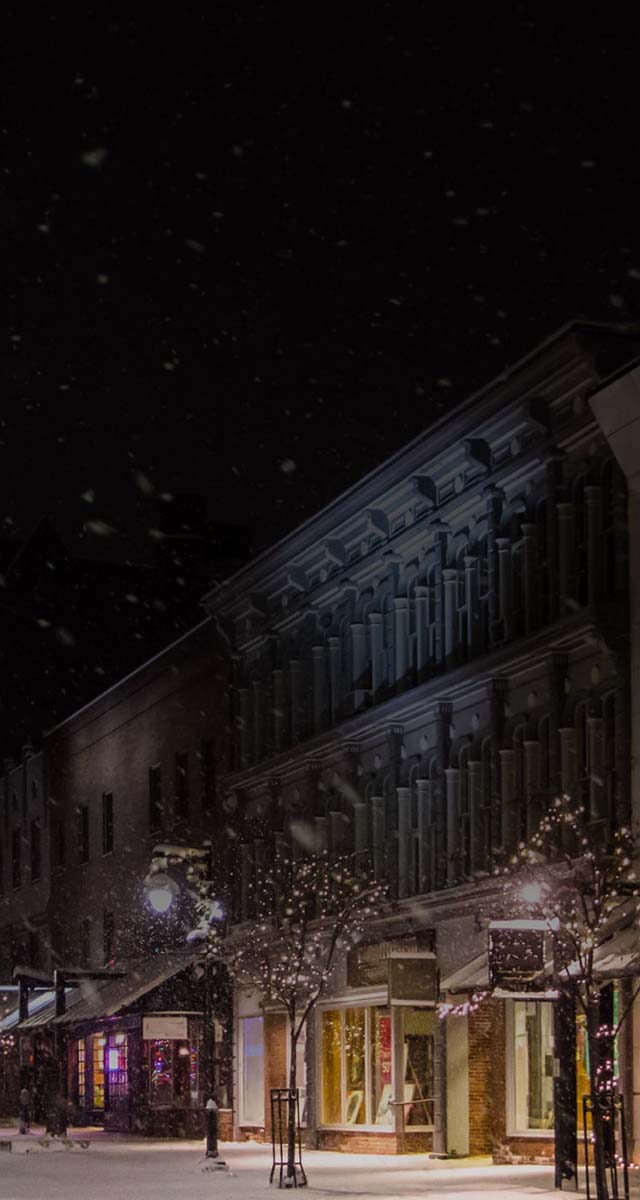 Photo of street with various storefronts at night with snow falling
